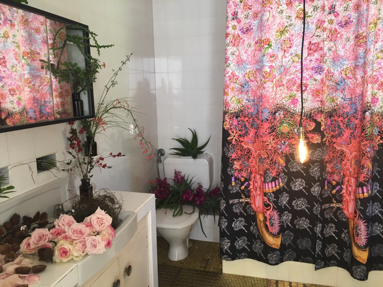 A bathroom with flower arrangements on the toilet and vanity and a detailed shower curtain displaying complex pattern of weapons and flowers.